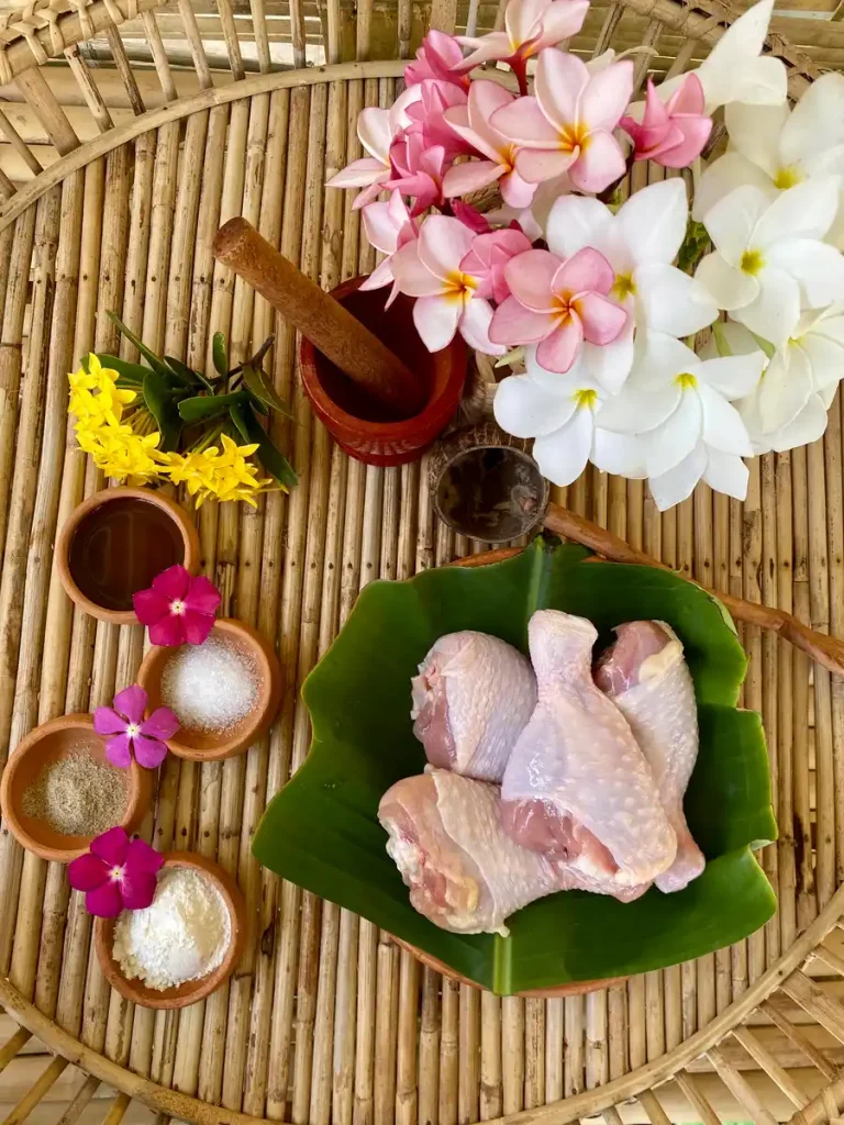 Raw chicken drumsticks, crispy frying flour, white sugar, fish sauce, and white pepper on a bamboo serving tray with several decorations such as flowers and a wooden spoon.