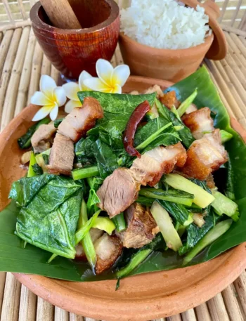 Clay dish of pad kana moo krob with white rice, a small mortar and pestle, and white flowers.