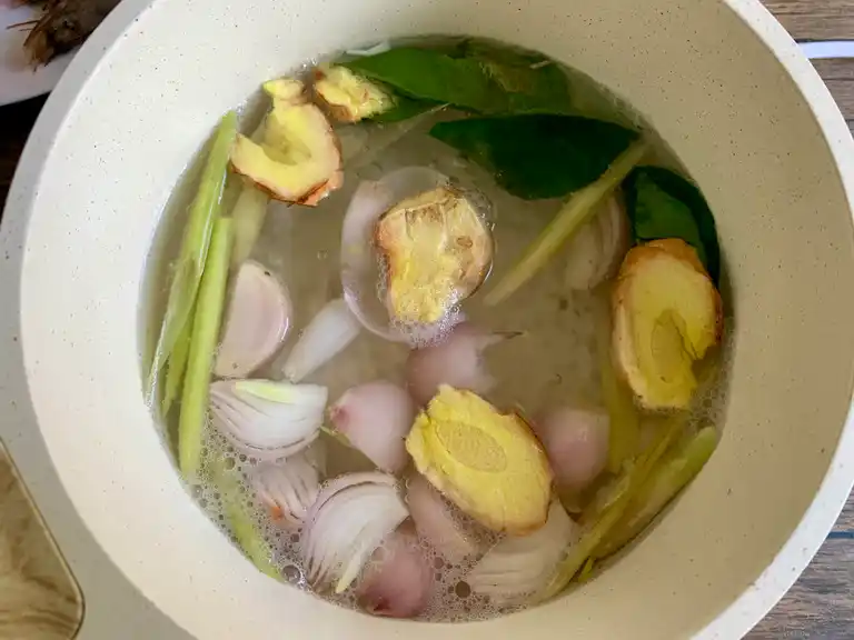 Shallots, galangal, lemongrass, and kaffir lime leaves simmering in a pot of water.