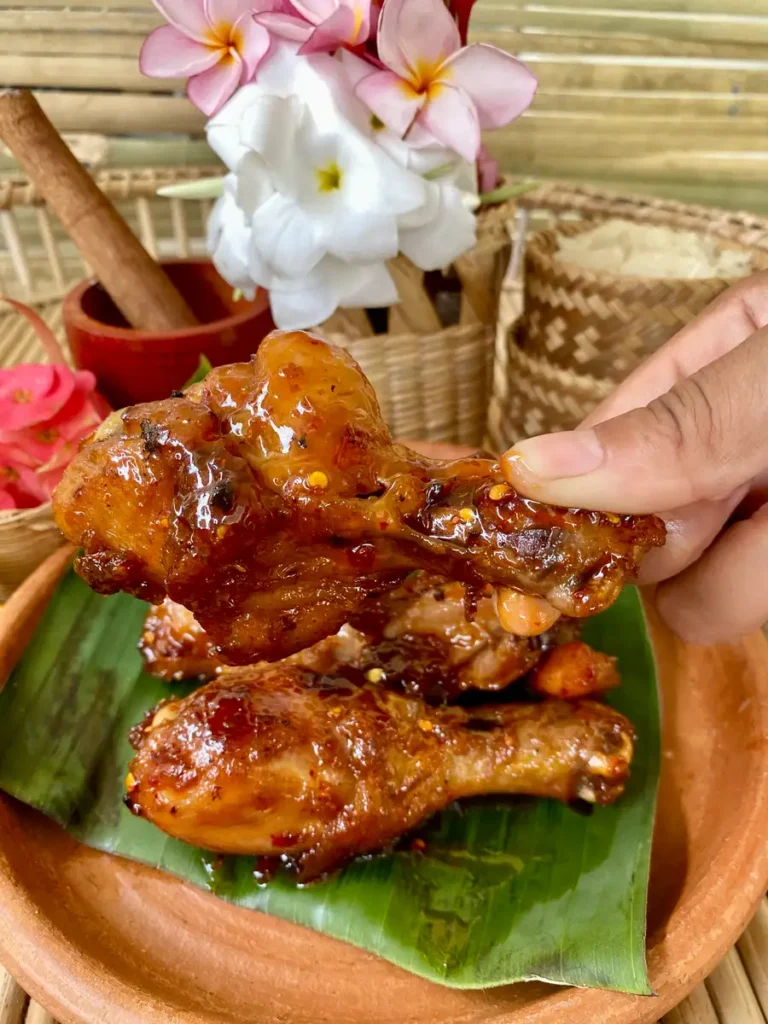 Hand lifting a sweet and spicy fried chicken drumstick over a clay dish with more fried chicken.