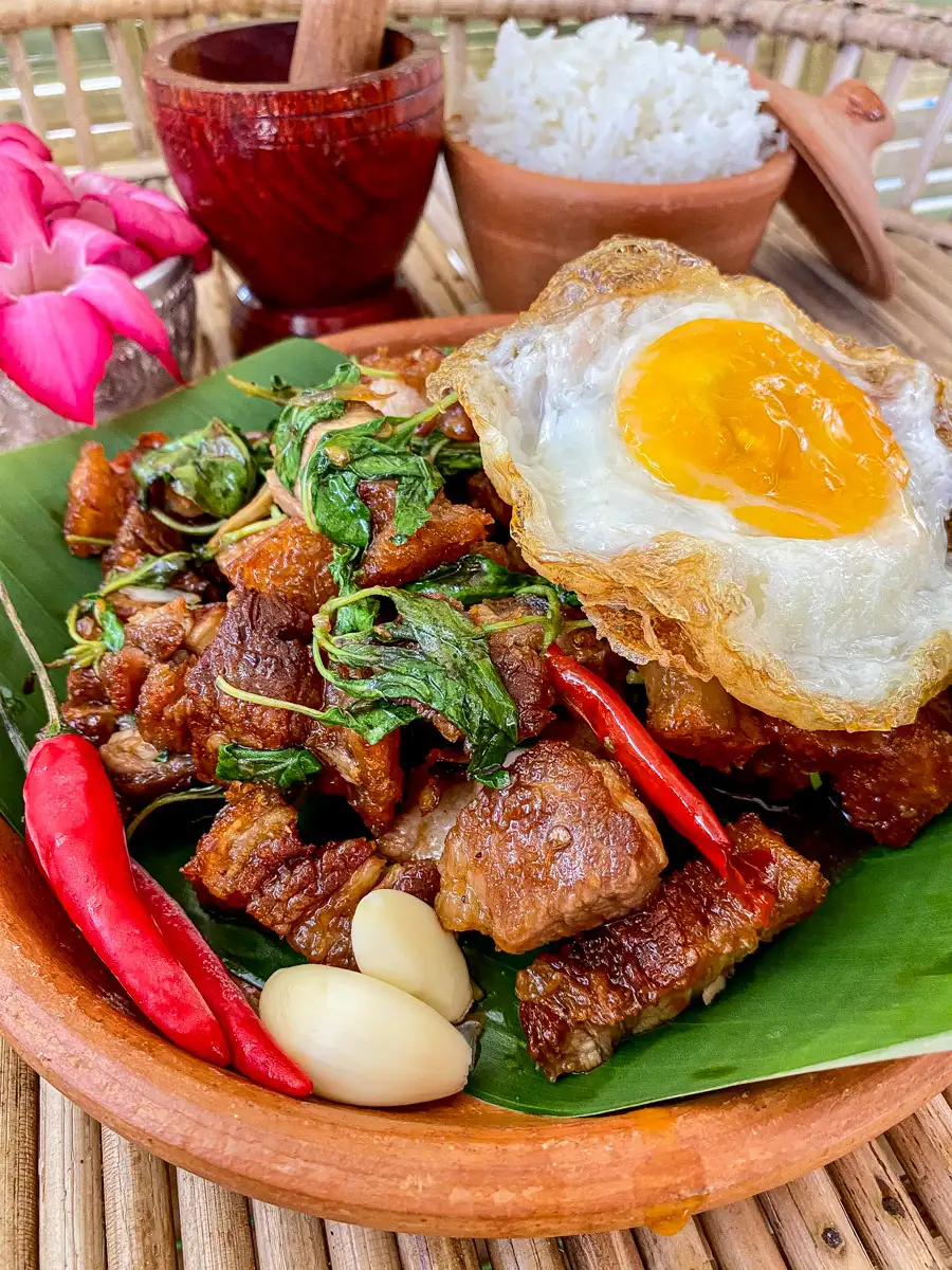 Thai basil pork belly with garlic, red chilies, and crispy fried egg served with rice in the background.