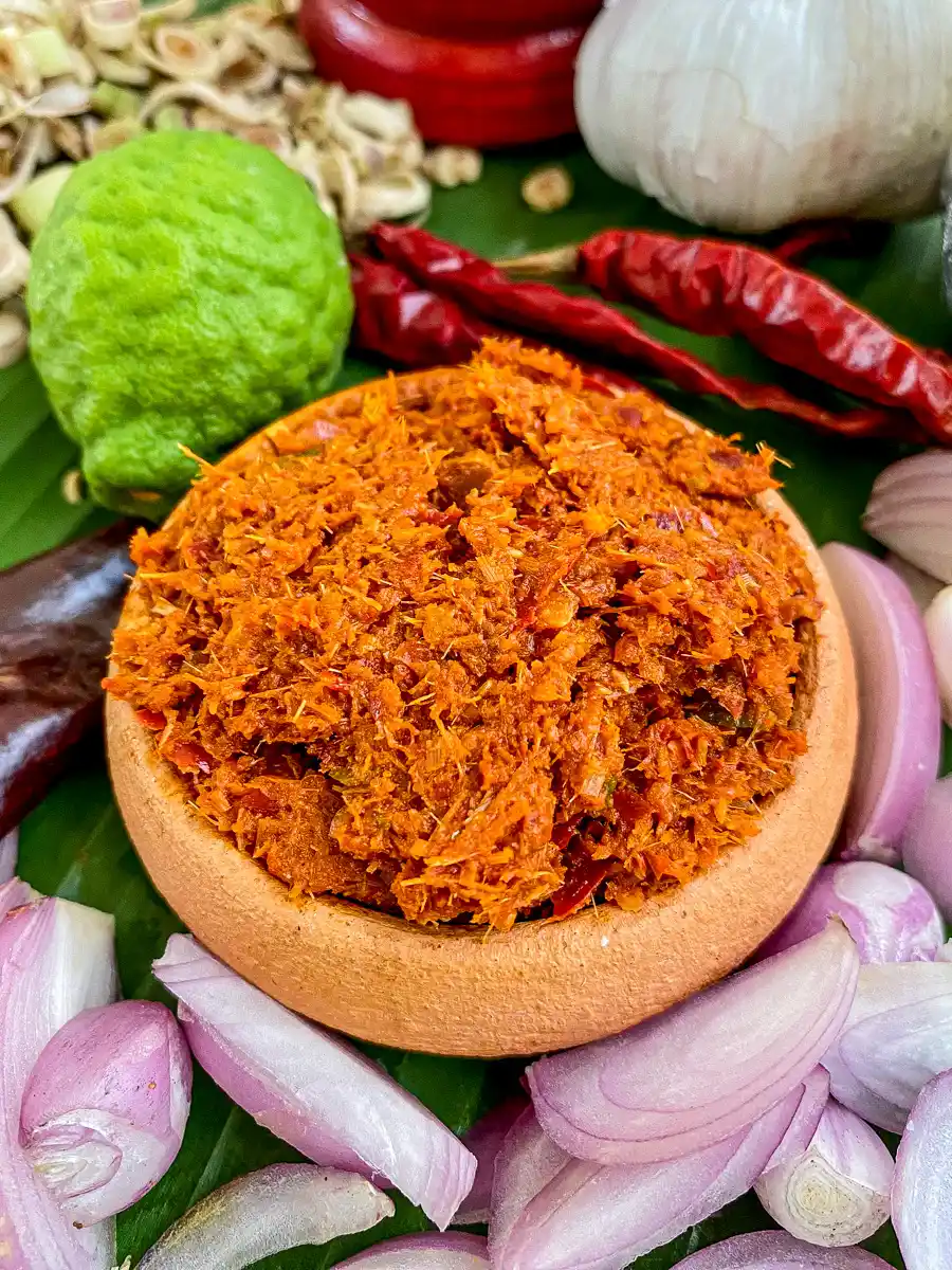 Thai red curry paste in a small clay bowl, with fresh lemongrass, garlic, shallots, and red chilies in the background.