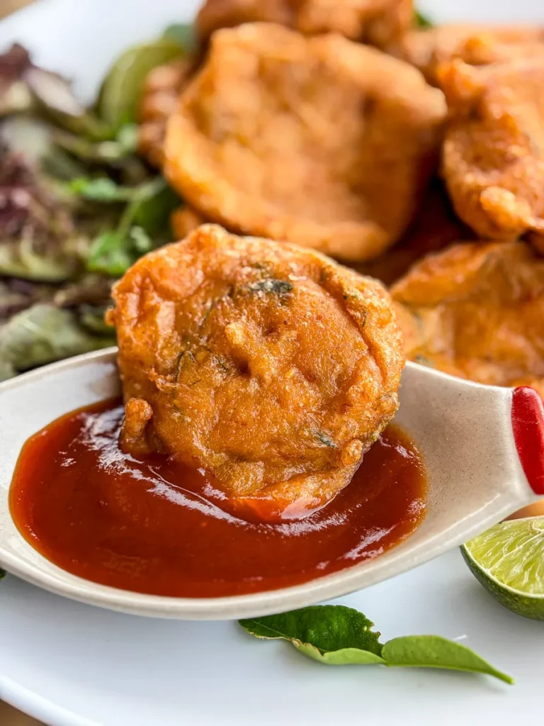 Close up of tod mun pla (Thai fish cake) dipped in sweet chili sauce, served with lime and herbs on the side.