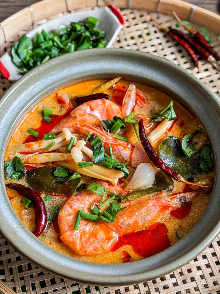 Tom yum kung soup in a clay bowl with shrimp, kaffir lime leaves, lemongrass, shallots, chili oil, and herbs.