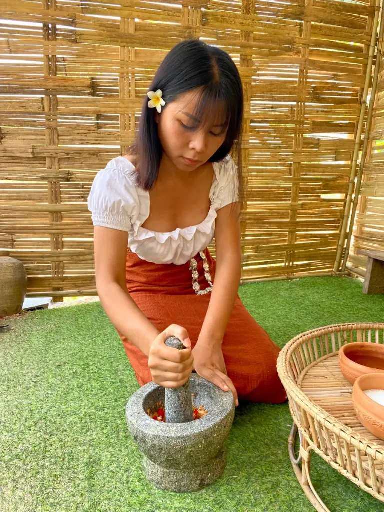 A Thai woman grinding red chilies and garlic in a granite mortar and pestle while kneeling on a grass floor.