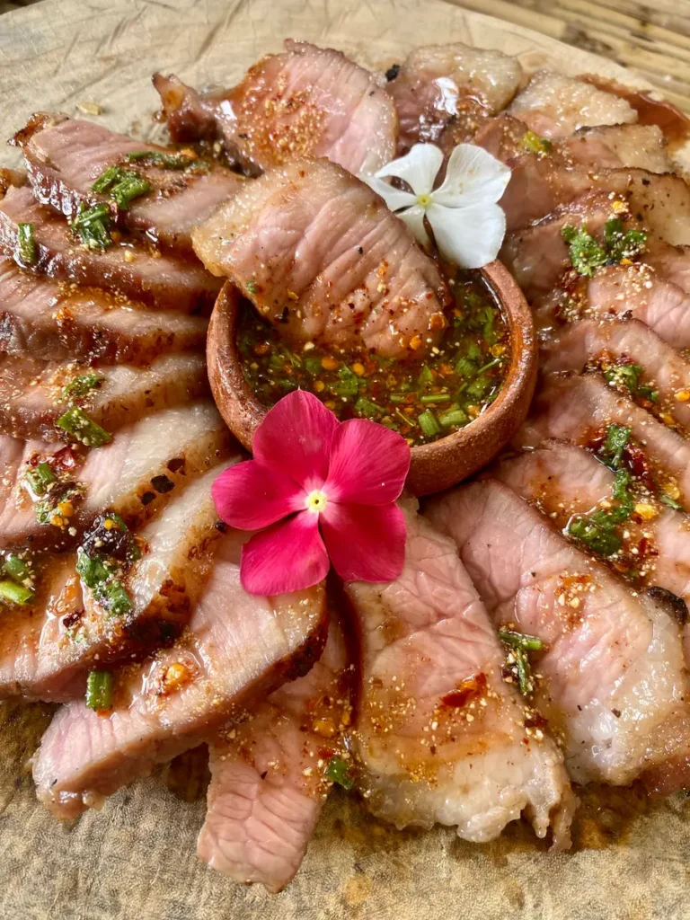 Sliced Thai crying tiger steak arranged on a wooden cutting board, drizzled with nam jim jaew dipping sauce, alongside a clay cup filled with spicy, tangy sauce at the center.