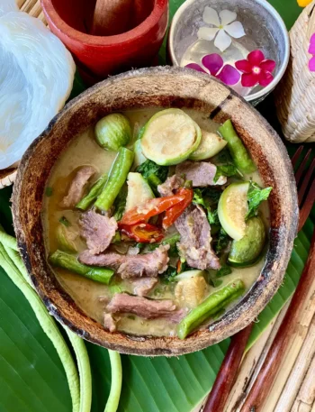 Authentic Thai green curry pork, known as gaeng keow wan, served in a coconut shell bowl with vegetables on a banana leaf.