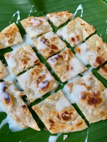 Thai roti dessert squares generously topped with sweetened condensed milk on a banana leaf.
