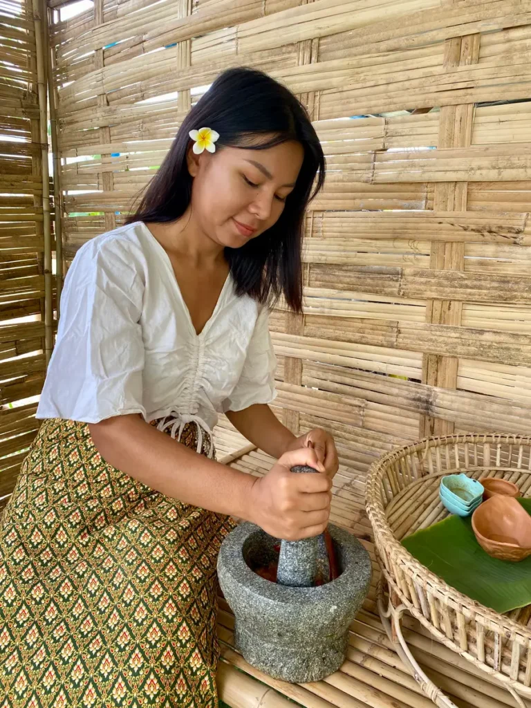 Thai woman seated on a bamboo table, working with a granite mortar and pestle.