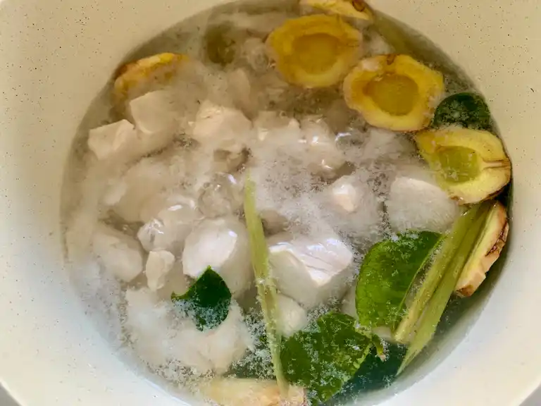 Sliced chicken breast simmering in a pot with galangal, lemongrass, and lime leaves.
