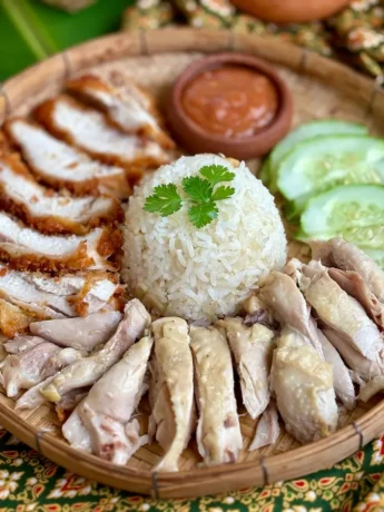 Khao man gai with chili sauce, accompanied by cucumber, steamed rice, and fried chicken.
