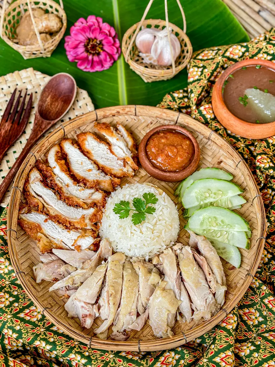 Thai chicken rice (khao man gai) with two styles of chicken, spicy dipping sauce, cucumber, and soup on a banana leaf setting.