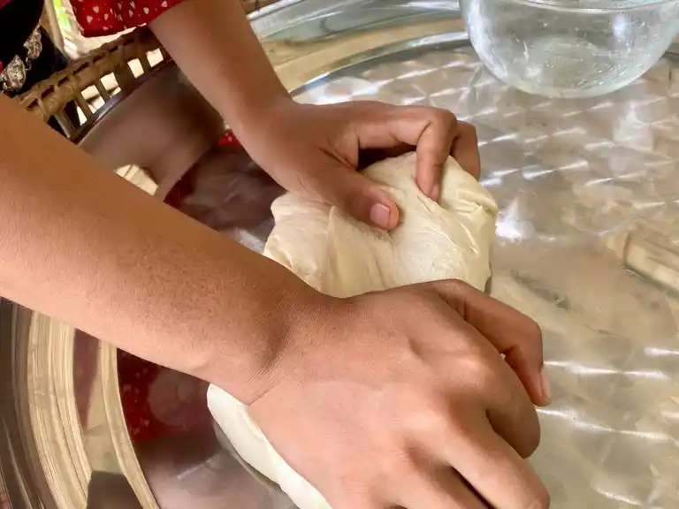 Hands kneading Thai roti dough on a round metal tray.