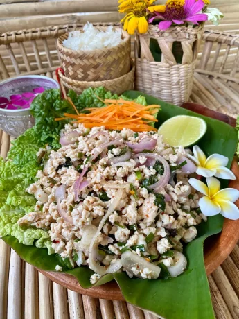 Larb gai salad served on a lettuce leaf, garnished with shredded carrot and a lime wedge, with a basket of sticky rice and decorative flowers in the background.