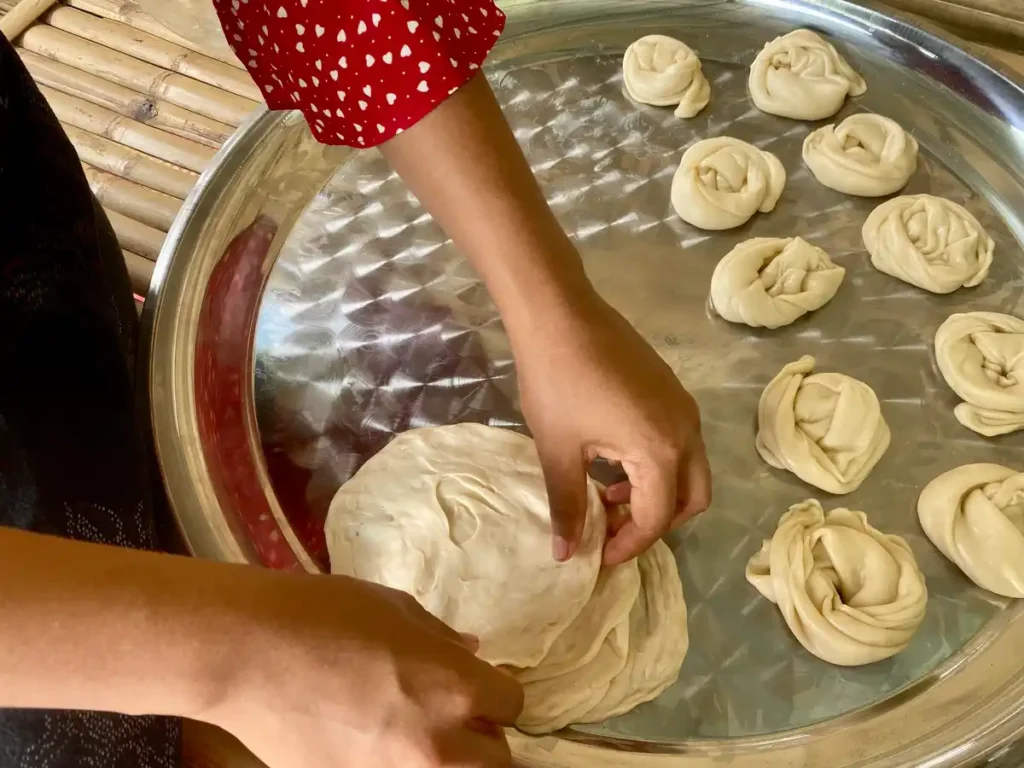 Stretching Thai roti dough by hand on a metal tray with prepped dough balls nearby.