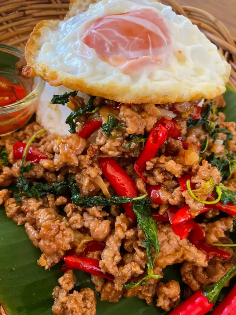 Authentic pad kra pao with minced pork and red chilies served over white rice, with a deep-fried egg. Served alongside a spicy chili sauce in a glass cup.