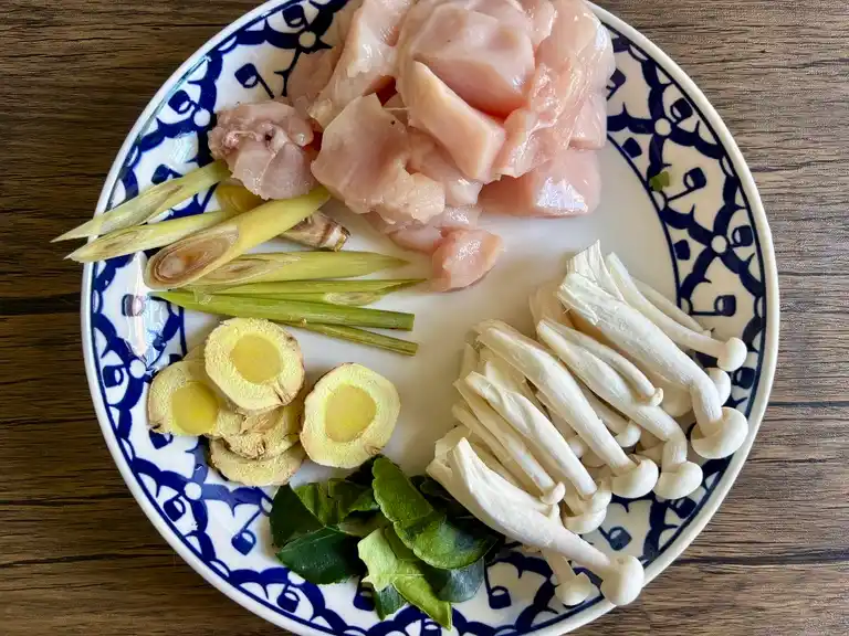 Sliced lemongrass, galangal, kaffir lime leaves, chicken breast, and enoki mushrooms on a blue and white Thai ceramic plate.