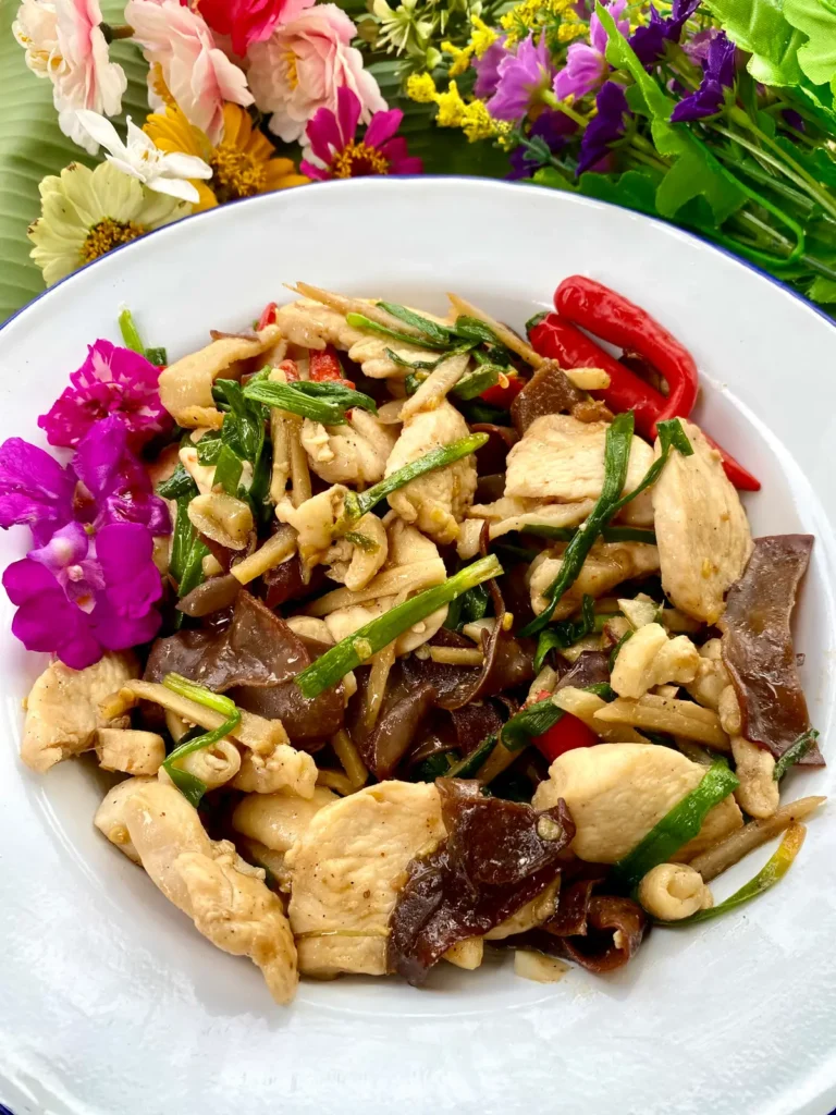 Thai ginger chicken in a white dish with red chilies, green onions, and cloud ear mushrooms. There are also decorative flowers.