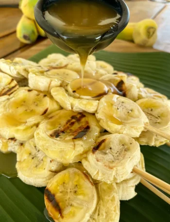 Grilled bananas on a skewer being drizzled with coconut caramel sauce that's in a black cup.