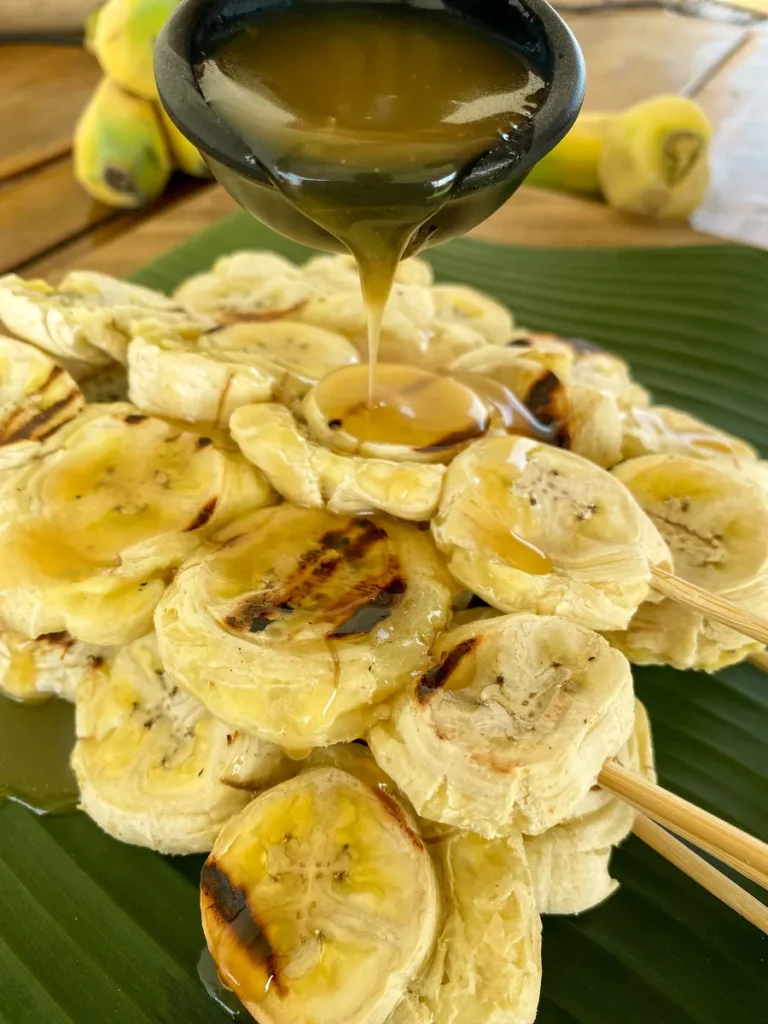 Grilled bananas on a skewer being drizzled with coconut caramel sauce that's in a black cup.