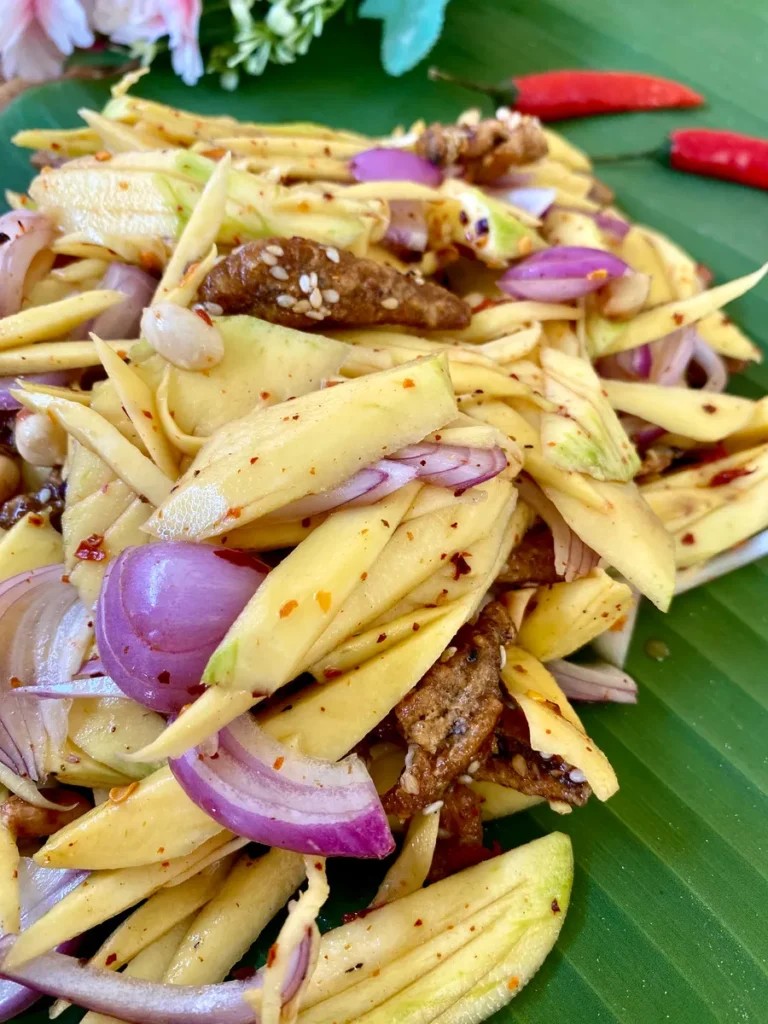 Close-up of raw Thai mango salad prepared with shallots, crispy fish snacks, and more traditional ingredients.