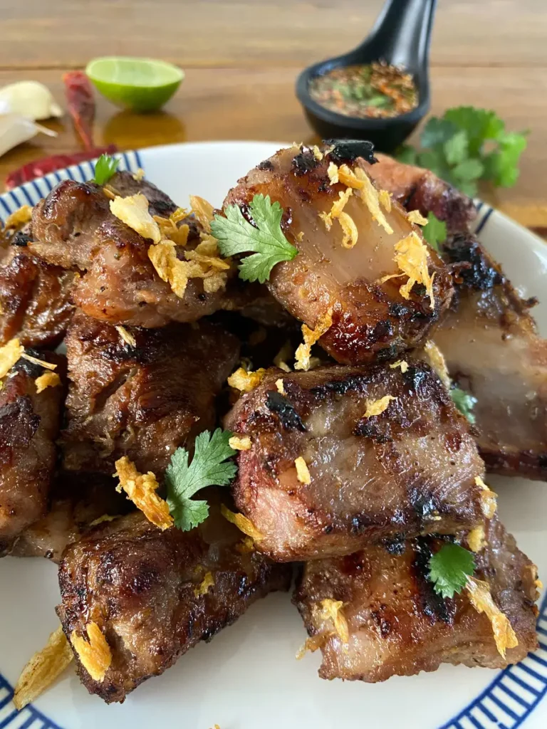 Grilled Thai ribs, garnished with coriander and crispy fried garlic, served on a white dish. In the background, a black cup with nam jim jaew chili dipping sauce, a lime wedge, dried chilies, garlic, and coriander.