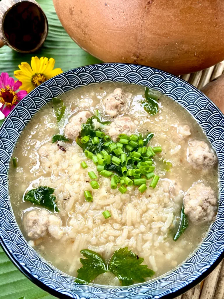 Thai rice soup, khao tom, with minced pork meatballs in a blue and white soup bowl, garnished with green onions and celery.