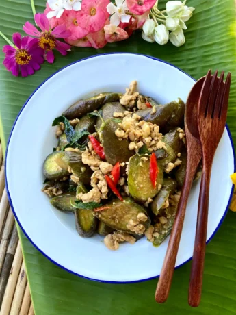Thai spicy eggplant stir-fry with minced chicken and chilies in a white dish, accompanied by a wooden fork and spoon. The dish is presented on top of a banana leaf that also has flowers at the top.