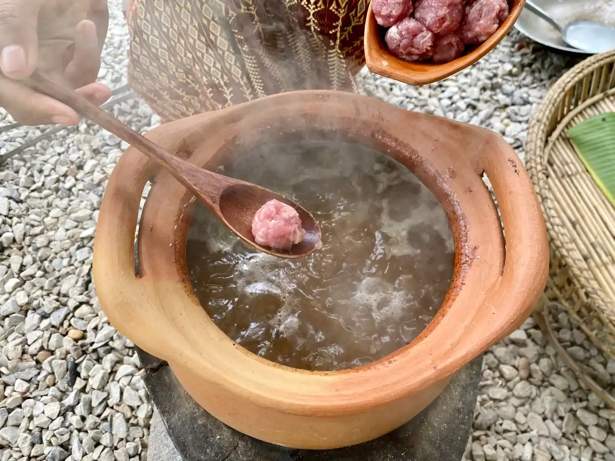 A clay soup pot with boiling broth, and a hand adding pork meatballs to it with the help of a wooden spoon.
