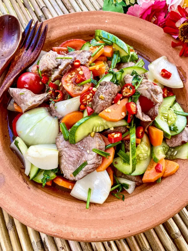 Authentic Thai beef salad served in a clay plate with cucumbers, carrots, onions, tomatoes, and fresh chili slices.