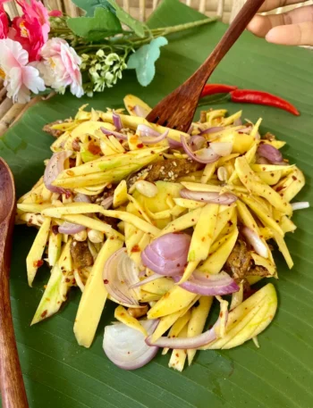 Authentic Thai mango salad featuring chili flakes, crispy fish snacks, and fine slices of shallots, with a wooden fork serving a portion.