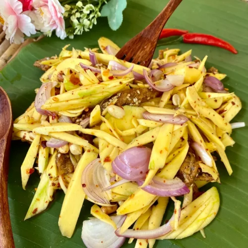 Authentic Thai mango salad featuring chili flakes, crispy fish snacks, and fine slices of shallots, with a wooden fork serving a portion.