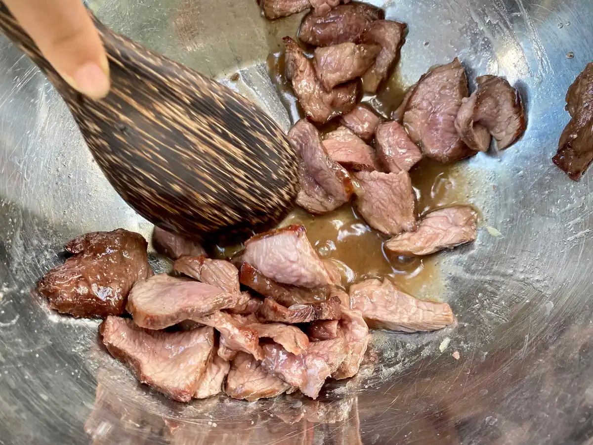 Close-up of a wooden spoon mixing beef slices with salad dressing in a mixing bowl.