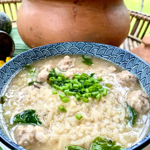 Khao tom, Thai rice soup with pork meatballs, in a blue-white soup bowl, garnished with green onions and celery. Behind the bowl is a large clay soup pot.
