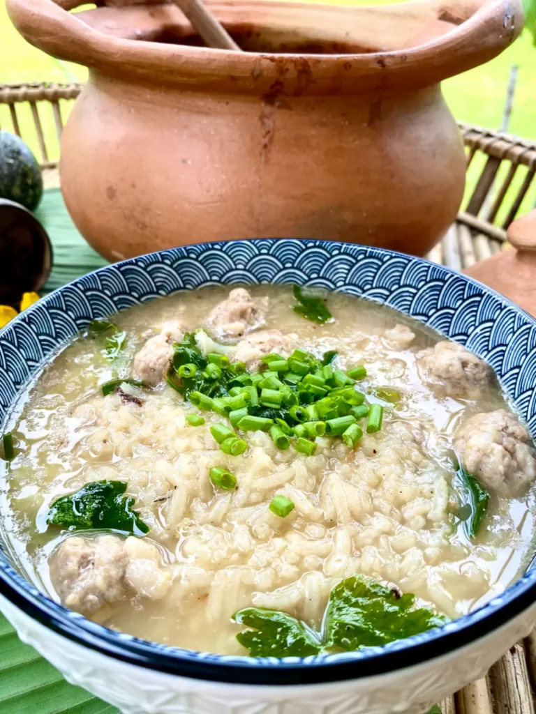 Khao tom, Thai rice soup with pork meatballs, in a blue-white soup bowl, garnished with green onions and celery. Behind the bowl is a large clay soup pot.