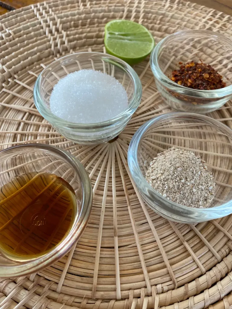 Ingredients for nam jim jaew sauce: white sugar, lime wedge, fish sauce, dried chili flakes, and toasted rice powder.