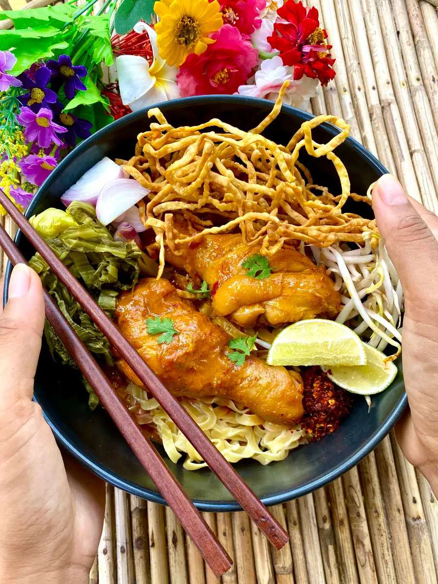 Authentic chicken khao soi noodle soup served with crispy noodles, pickled mustard greens, lime, and chili flakes in a black bowl.