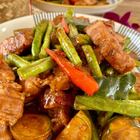 Pad prik king with pork, green beans, kaffir lime leaves, and Thai eggplants in a white dish. Behind it is another blurred out portion of the same dish.