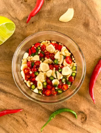 Top-down view of prik nam pla prepared in a clear glass cup, encircled by fresh red and green chilies, garlic, and fresh lime segments.
