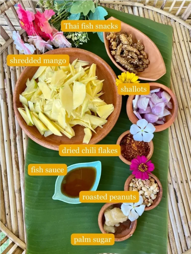 Shredded mango, Thai fish snacks, sliced shallots, dried chili flakes, fish sauce, roasted peanuts, and palm sugar presented in clay cups on a banana leaf.