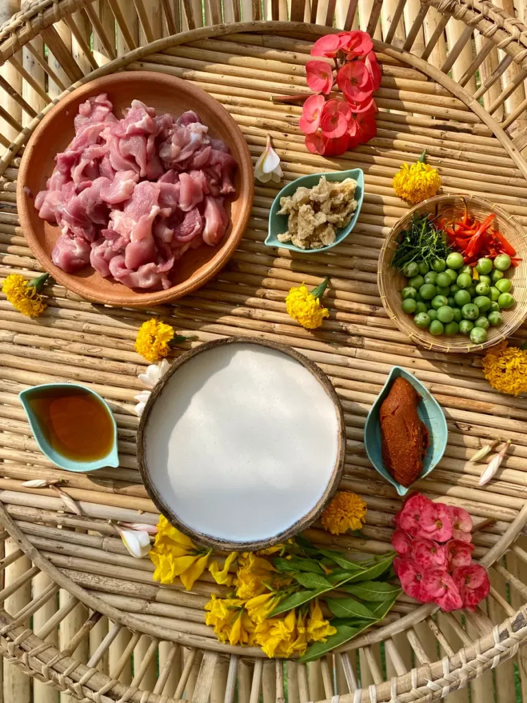 Ingredients to make this recipe on a bamboo serving tray: Coconut milk in a coconut shell, panang curry paste, palm sugar, and fish sauce in plastic cups. Snow peas, chilies, and lime leaves in a bamboo cup, and sliced pork in a clay dish.