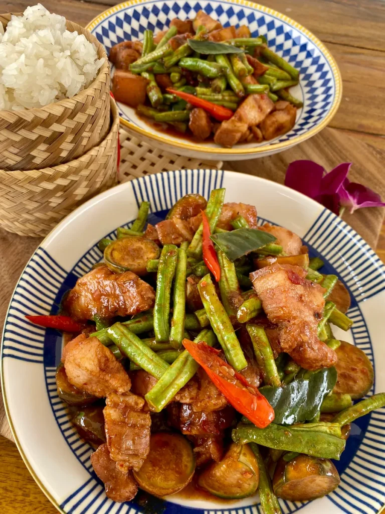 2 Portions of red curry stir-fry with green beans in a white dish, and a portion of sticky rice in a bamboo rice pot.