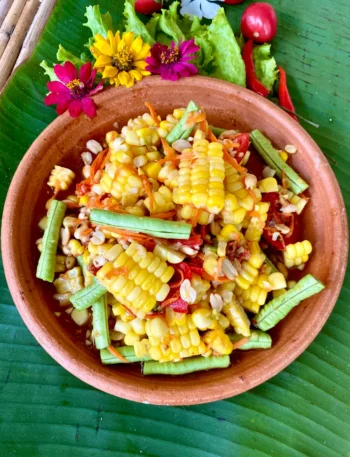 Thai corn salad made with sweetcorn, yard-long beans, tomatoes, red chilies, and carrots, served in a clay dish on a banana leaf.