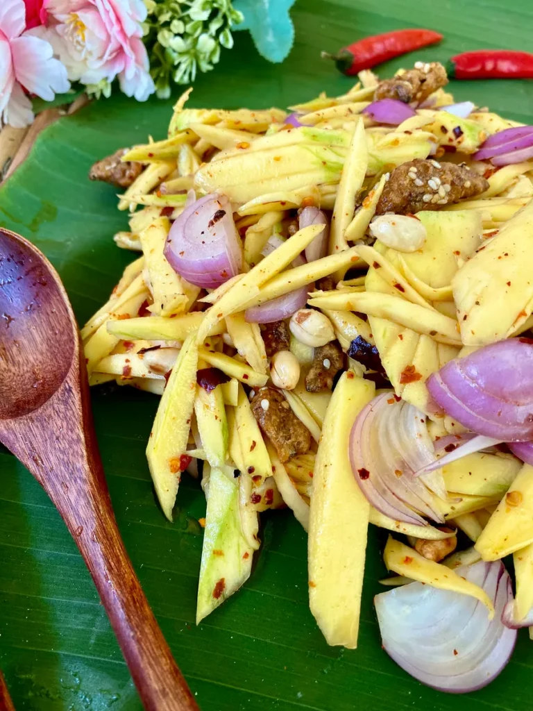 Traditional Thai mango salad coated with spices, presented on a banana leaf with a wooden spoon.