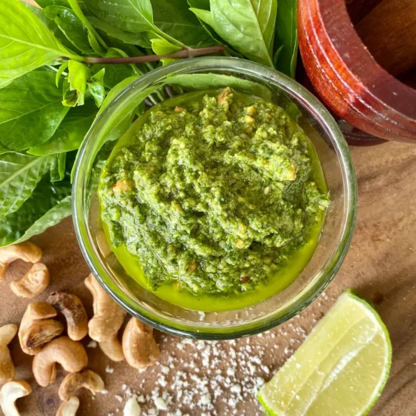 Top-down view of Thai basil pesto in a glass cup on a wooden cutting board. Scattered around it are roasted cashews, a lime wedge, salt, Thai basil, and a small wooden mortar and pestle.