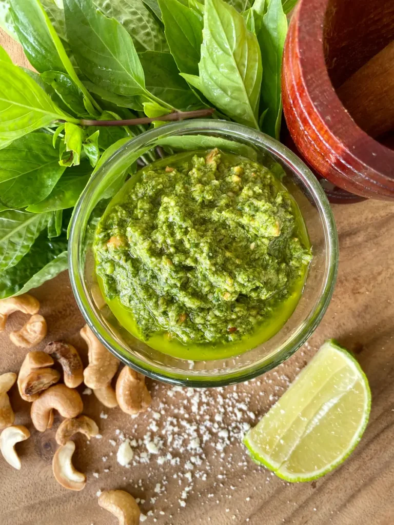 Top-down view of Thai basil pesto in a glass cup on a wooden cutting board. Scattered around it are roasted cashews, a lime wedge, salt, Thai basil, and a small wooden mortar and pestle.