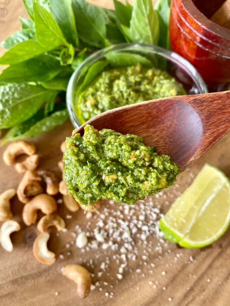 A wooden spoon with Thai basil pesto over a glass cup with more of the sauce. There's also basil leaves, cashews, salt, and a lime wedge on a wooden cutting board.