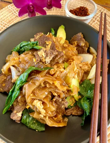 Thai beef pad see ew in a clay dish with Chinese broccoli, onions, and garlic. On the plate is a pair of wooden chopsticks, and next to it is a glass cup with red chili flakes.