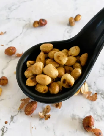 Dry roasted peanuts in a black spoon on a white background, with more nuts spread around the spoon.