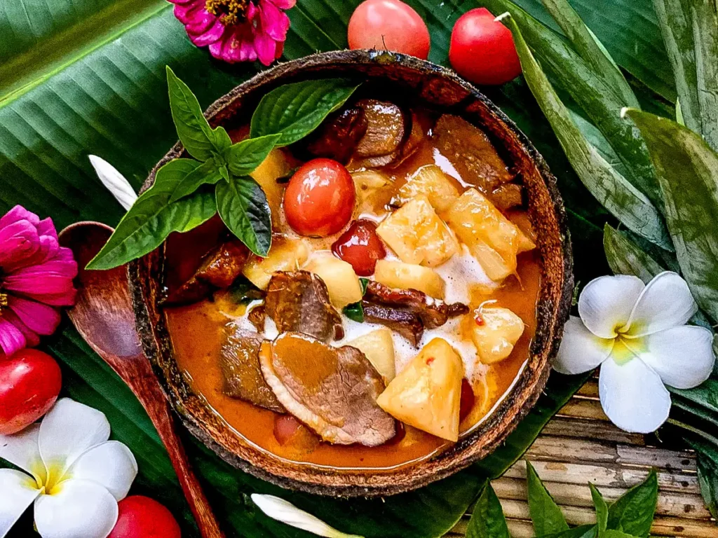 Gaeng phed ped yang, a Thai duck curry with coconut milk, pineapple, and cherry tomatoes, served in a coconut bowl and garnished with sweet basil.